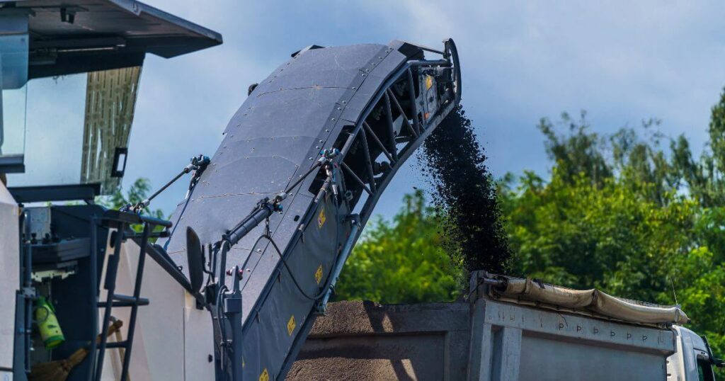 A machine performing asphalt milling services, grinding old asphalt and loading it into a truck for removal.